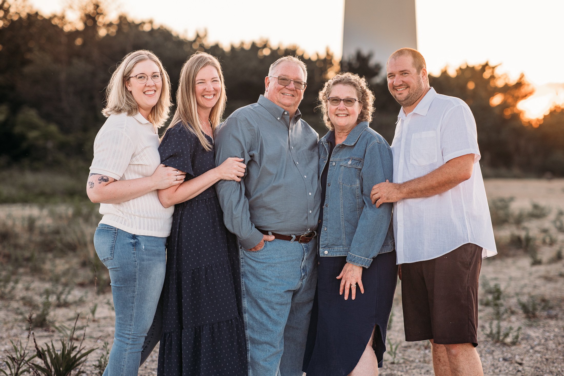 A family of six standing outdoors at sunset, smiling, with a lighthouse in the background.
