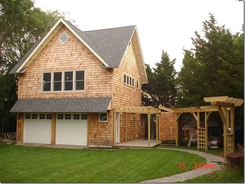 A two-story house under construction with wooden siding, a two-car garage, and an unfinished wooden pergola in the backyard, with greenery and trees in the background.