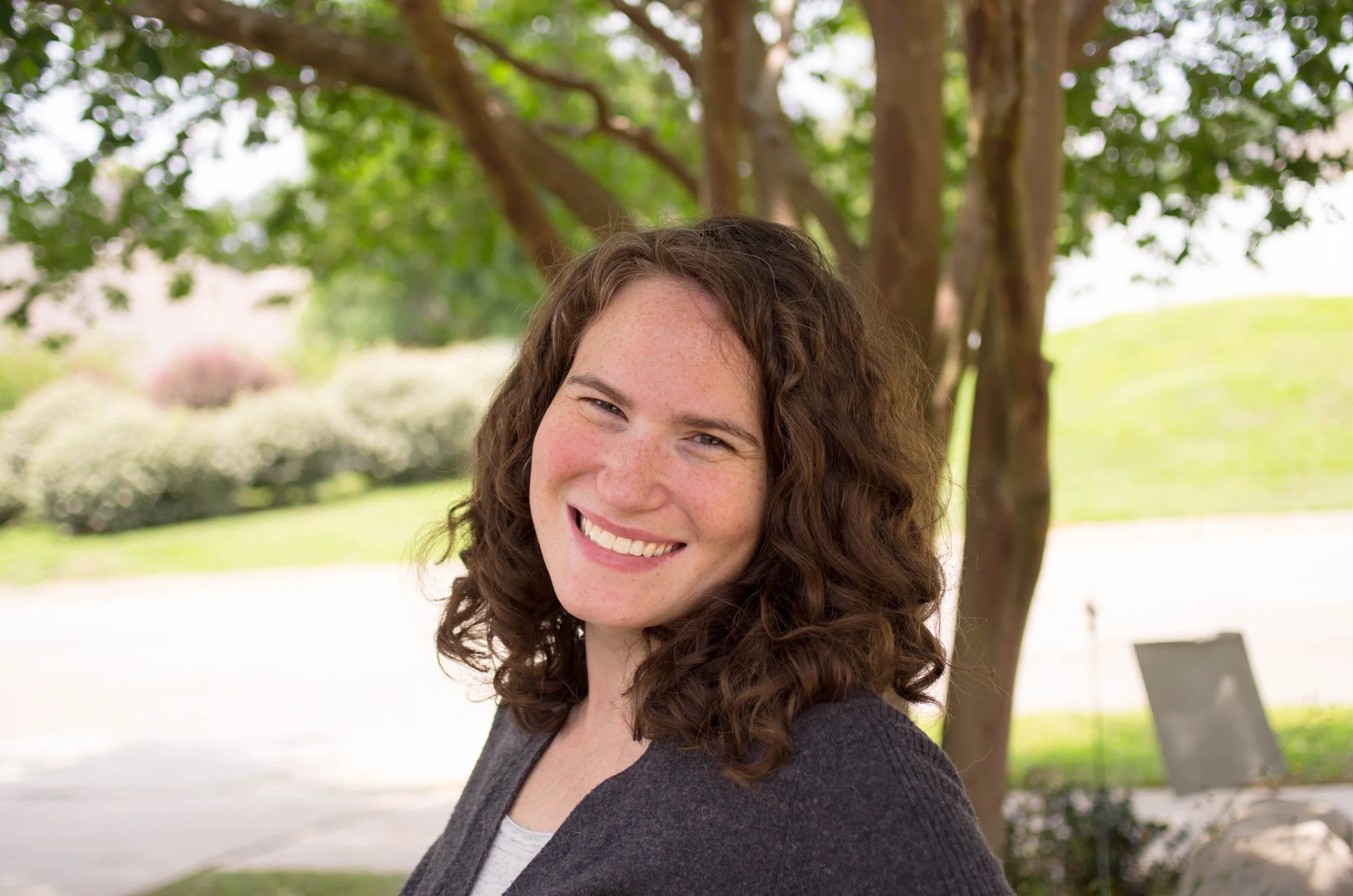 A smiling woman with curly brown hair and a gray cardigan standing outdoors near a large tree, with greenery and a view of a park or yard in the background.