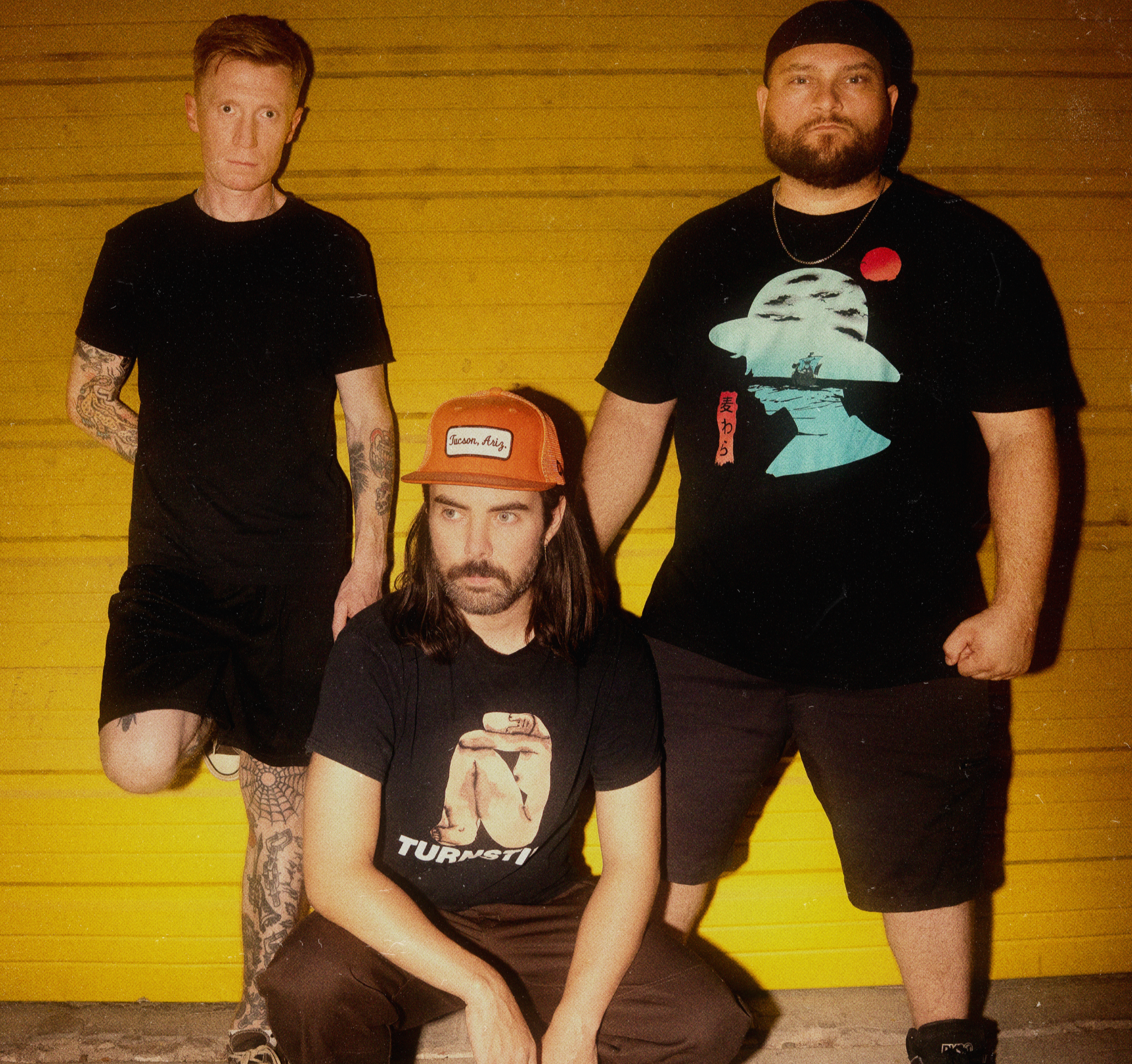 Three men posing against a yellow wooden wall, one seated in front, one standing on the left, and one on the right, dressed casually with graphic T-shirts and tattoos.