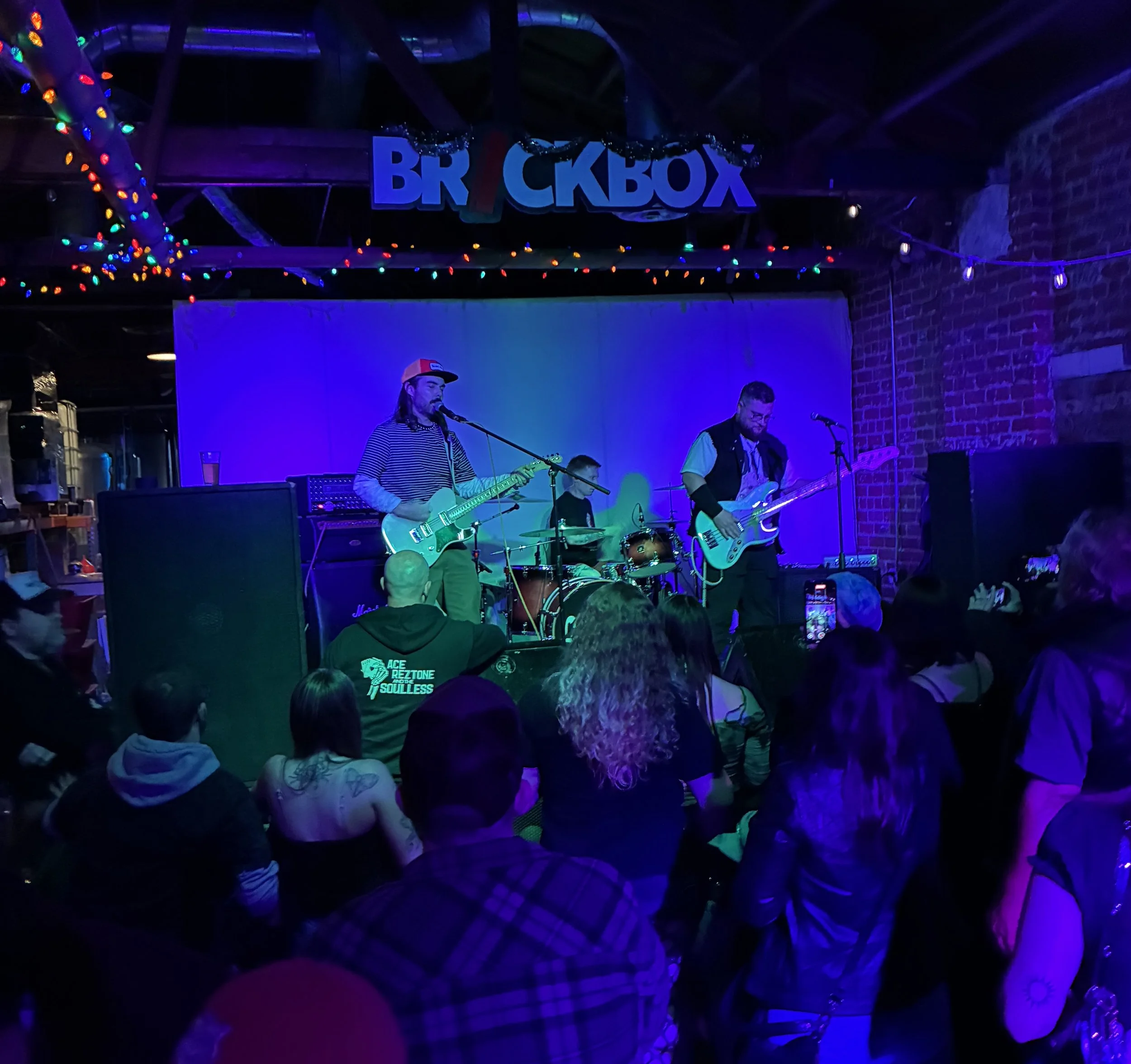 Live music performance on stage at a venue called 'Brickbox', with band playing guitar and drums, audience watching in a dimly lit space with colorful stage lighting and string lights overhead.