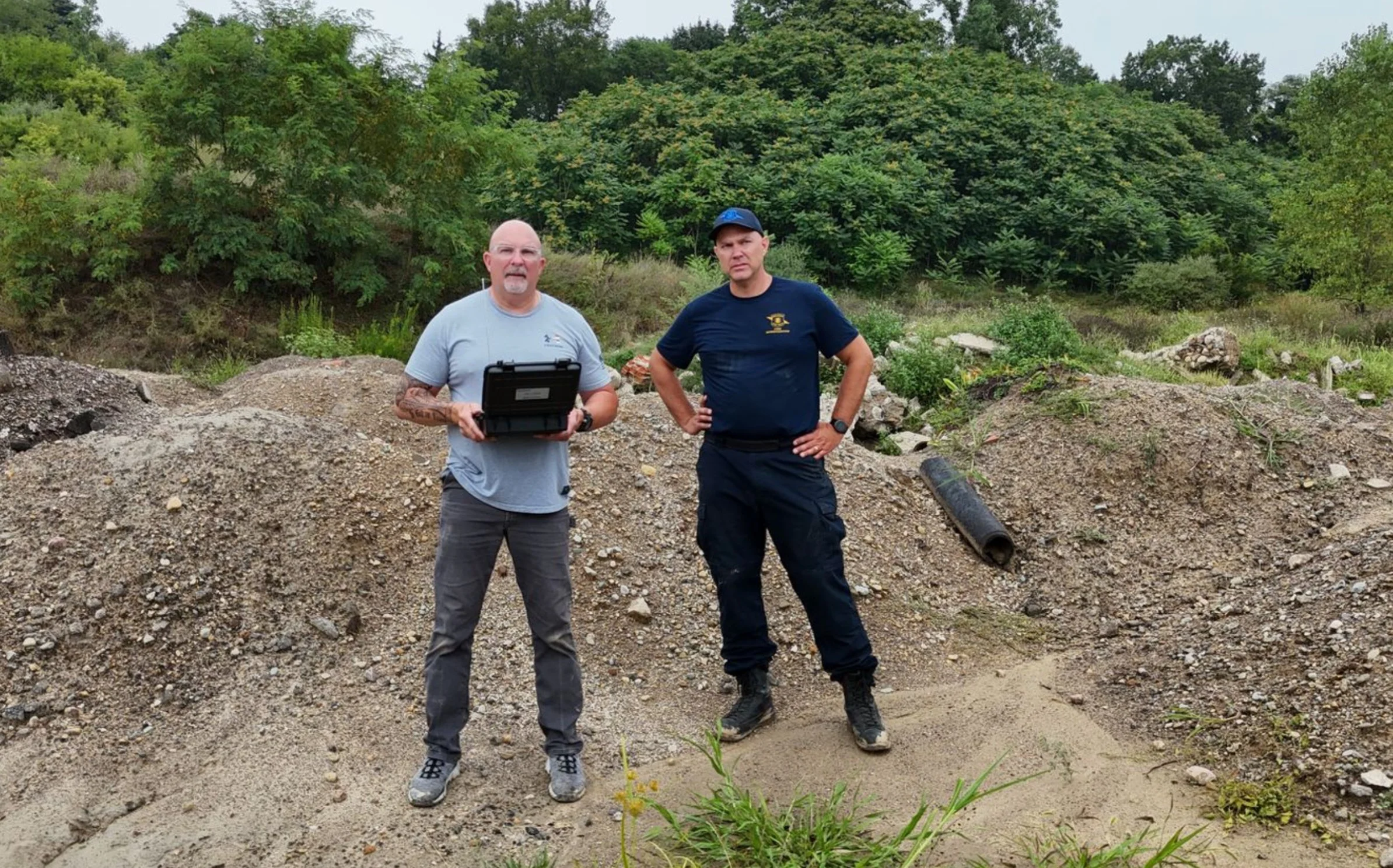 Two men standing outdoors on a dirt mound with trees and green foliage in the background. One man is holding a black case, and both are dressed in casual and tactical clothing.