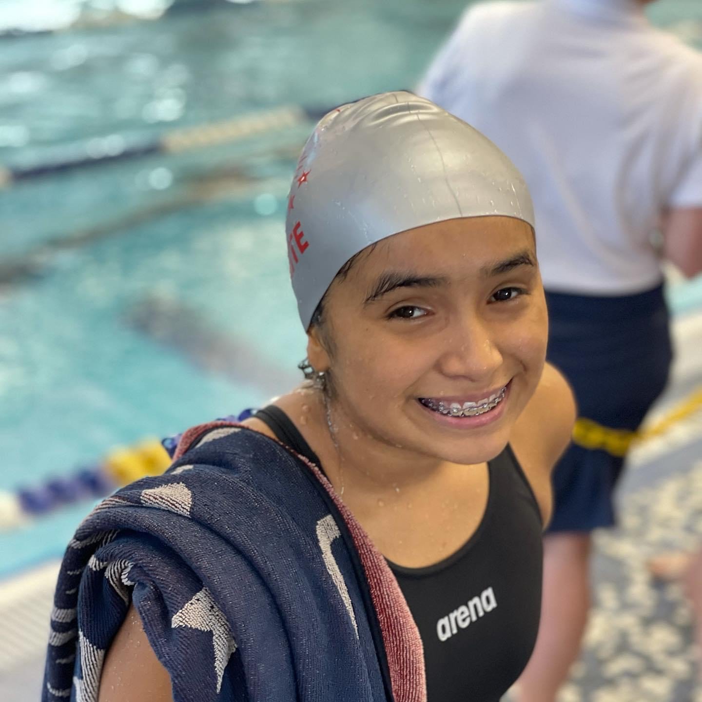 Young female swimmer with a gray swim cap and braces smiling at the camera after swimming practice, with a towel over her shoulder in a swimming pool.