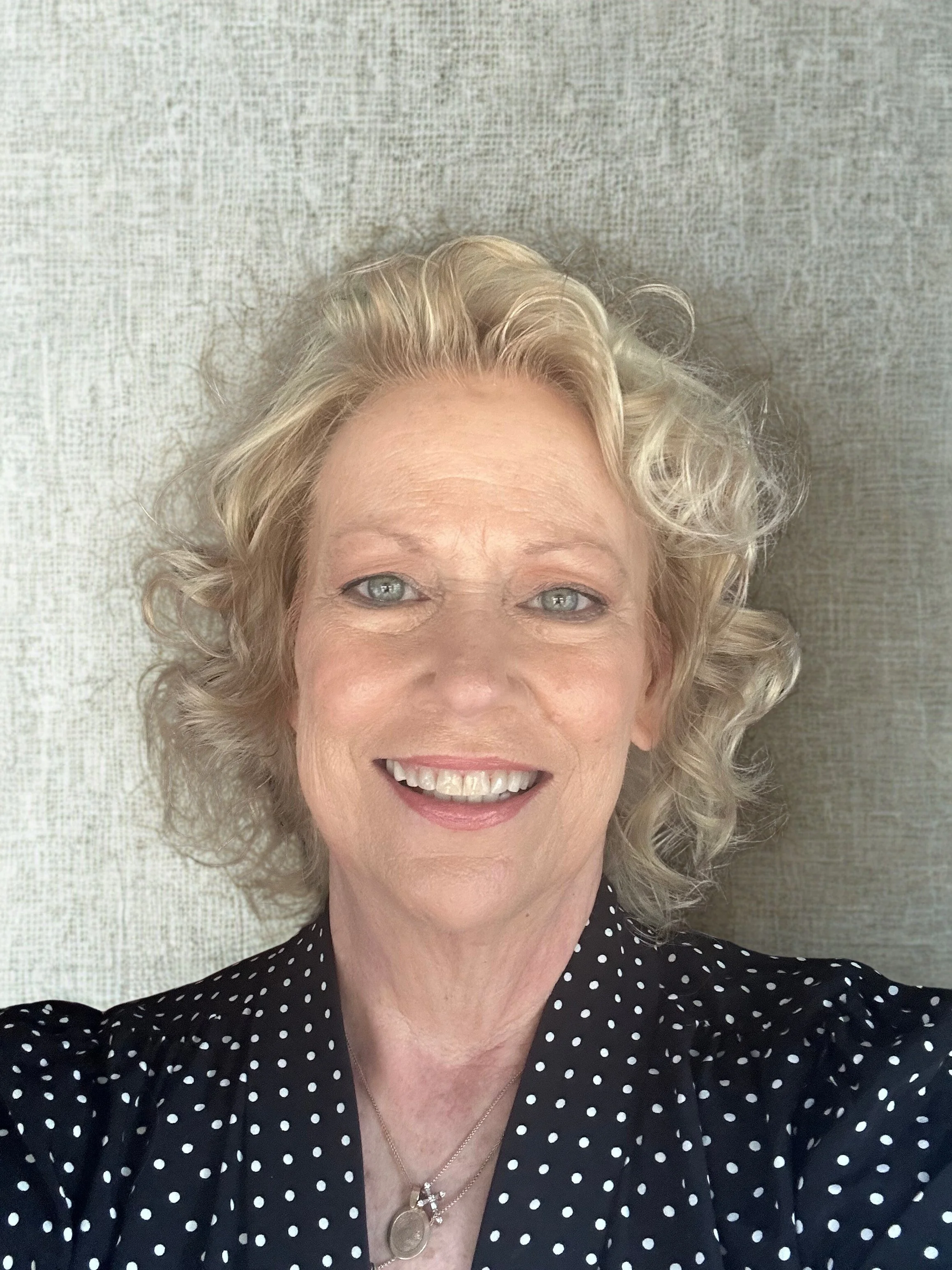 A smiling woman with curly blond hair, blue eyes, wearing a black dress with white polka dots and a silver necklace.