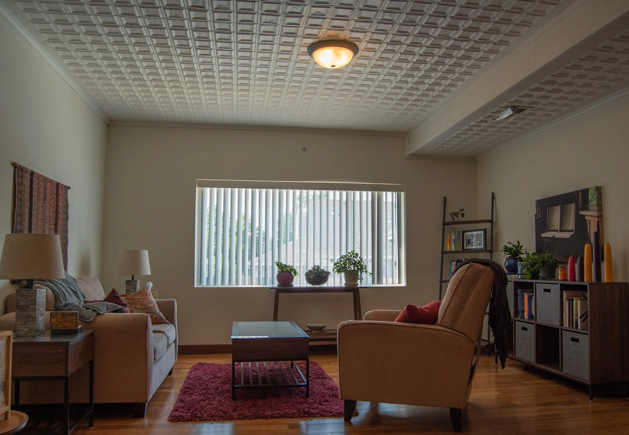 The view of therapy space in Janet's office with beige sofa, armchair, coffee table, and window with vertical blinds. Decor includes potted plants, picture frames, and candles, with a ceiling light and wood flooring.