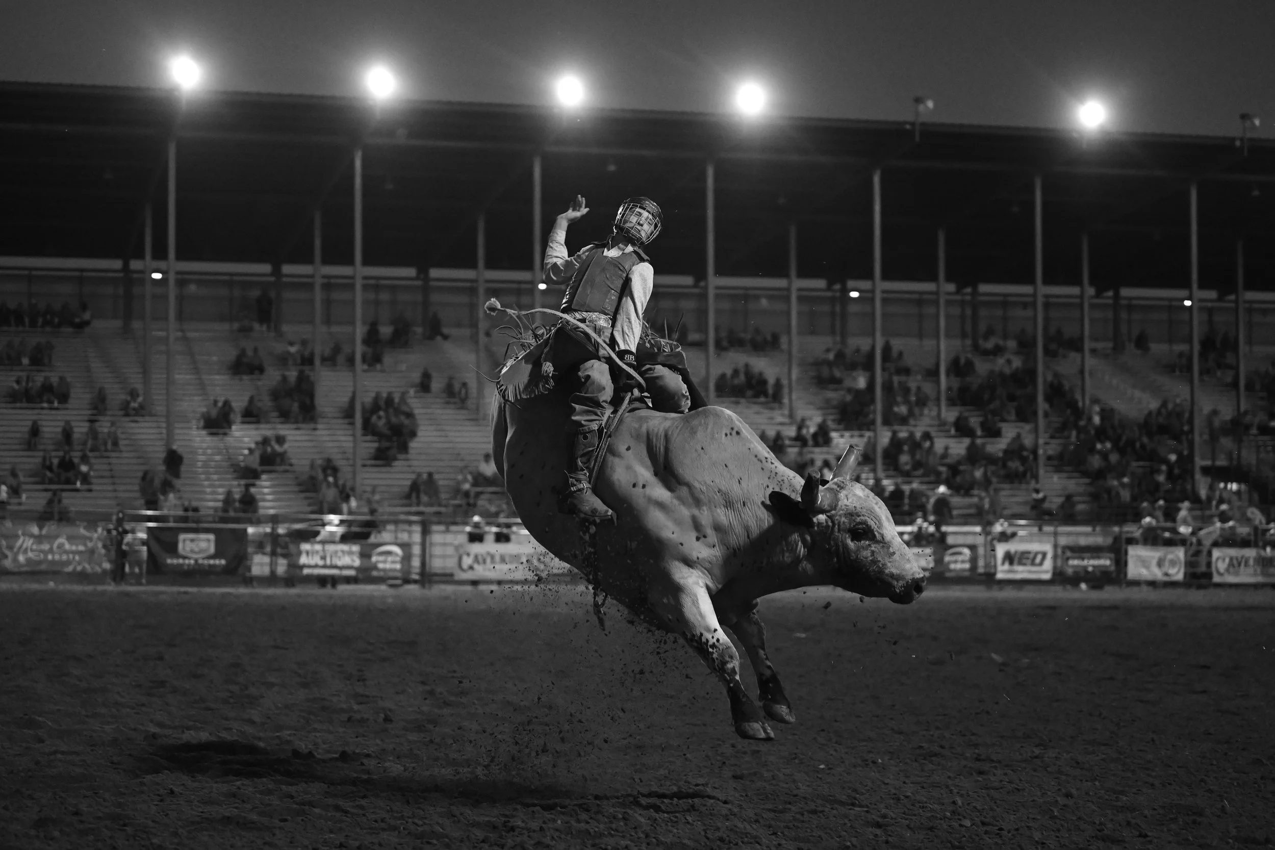 A rodeo cowboy in a helmet and protective vest riding a bucking bull at night, with a crowd sitting in the stands in the background.