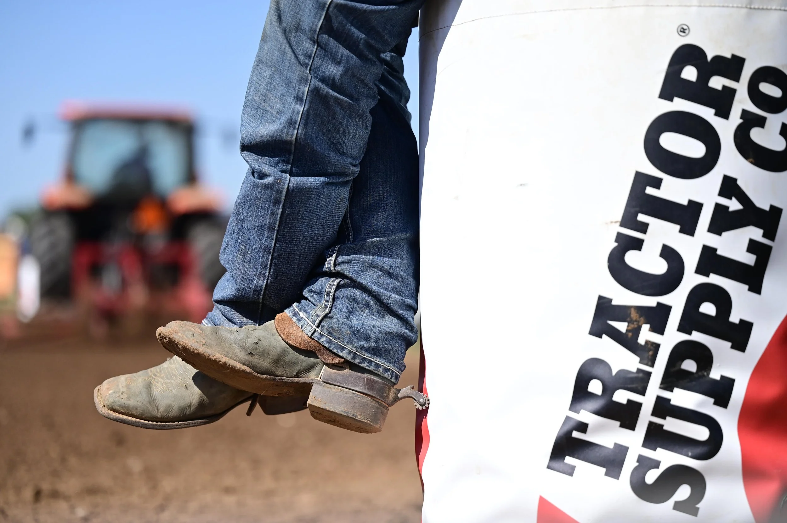 Close-up of a cowboy's legs with jeans, cowboy boots, and spurs, sitting on a barrel in a rodeo arena with a tractor driving away in the background.
