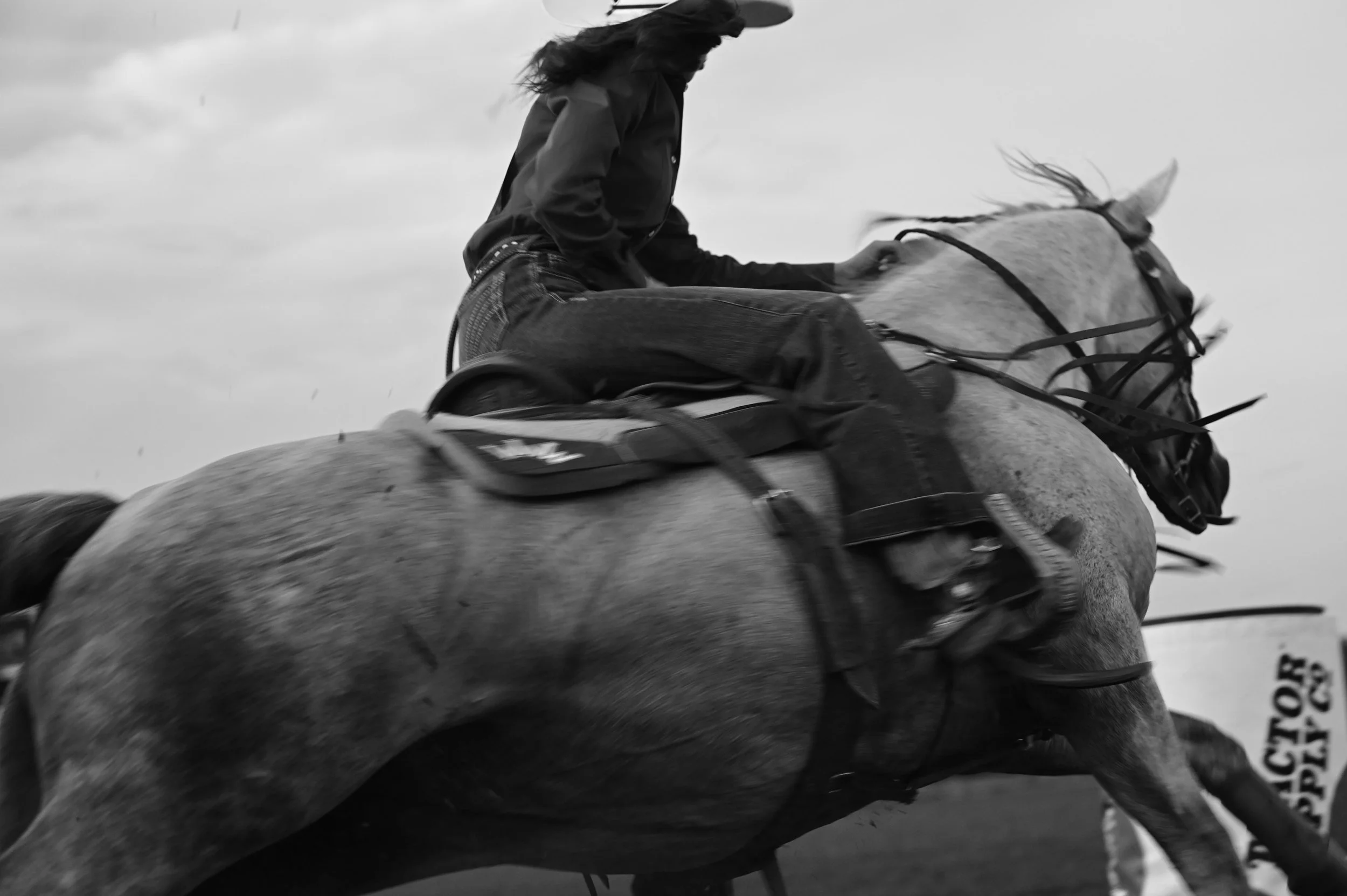 A barrel racer riding a horse around a barrel at a rodeo, captured in motion in black and white.
