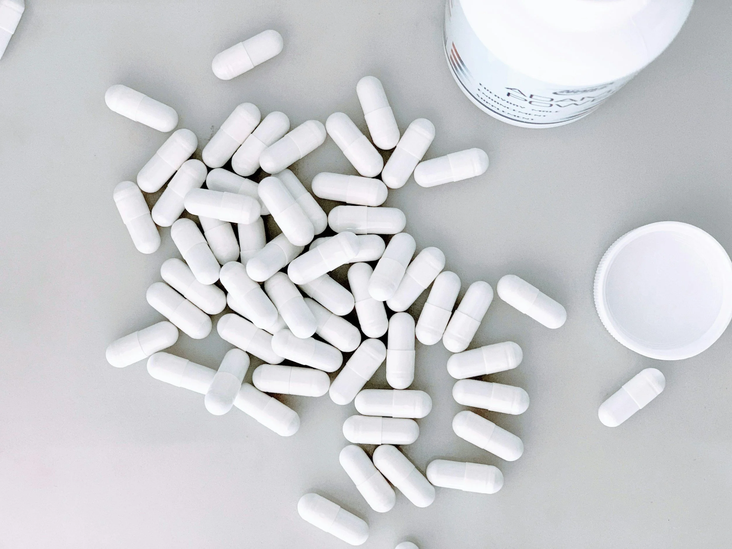White capsules spilled on a light gray surface with a white pill bottle and its open cap in the background.