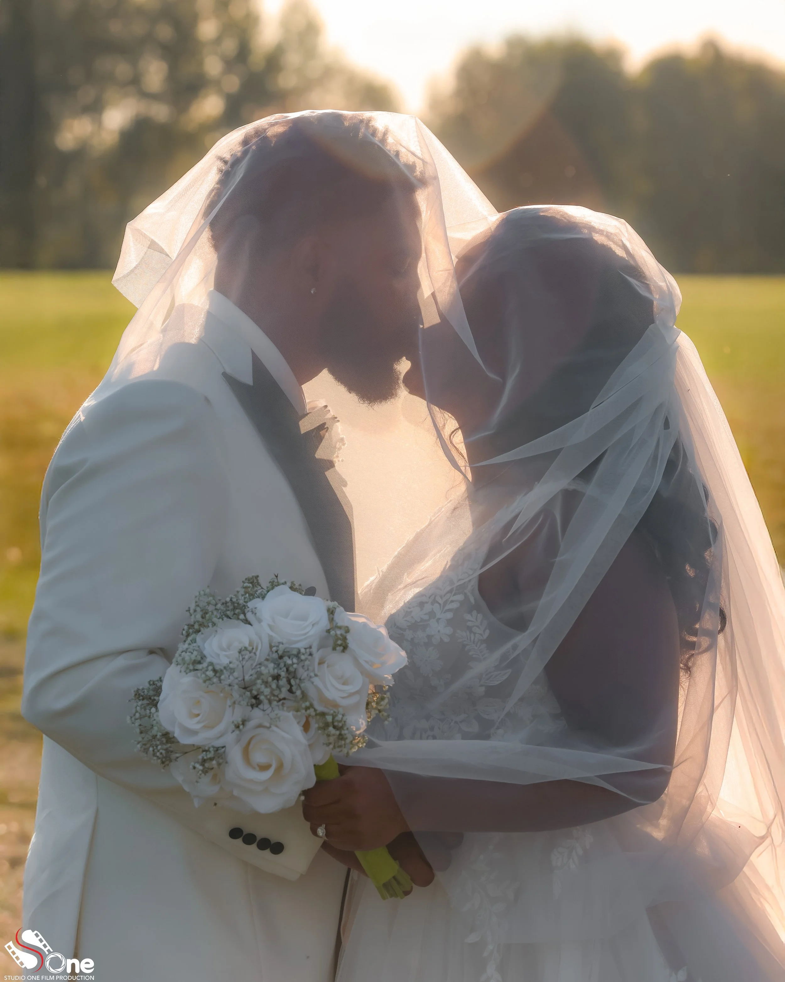 A bride and groom are touching foreheads, wearing wedding attire and veils, with a bouquet of white roses, standing outdoors at sunset.