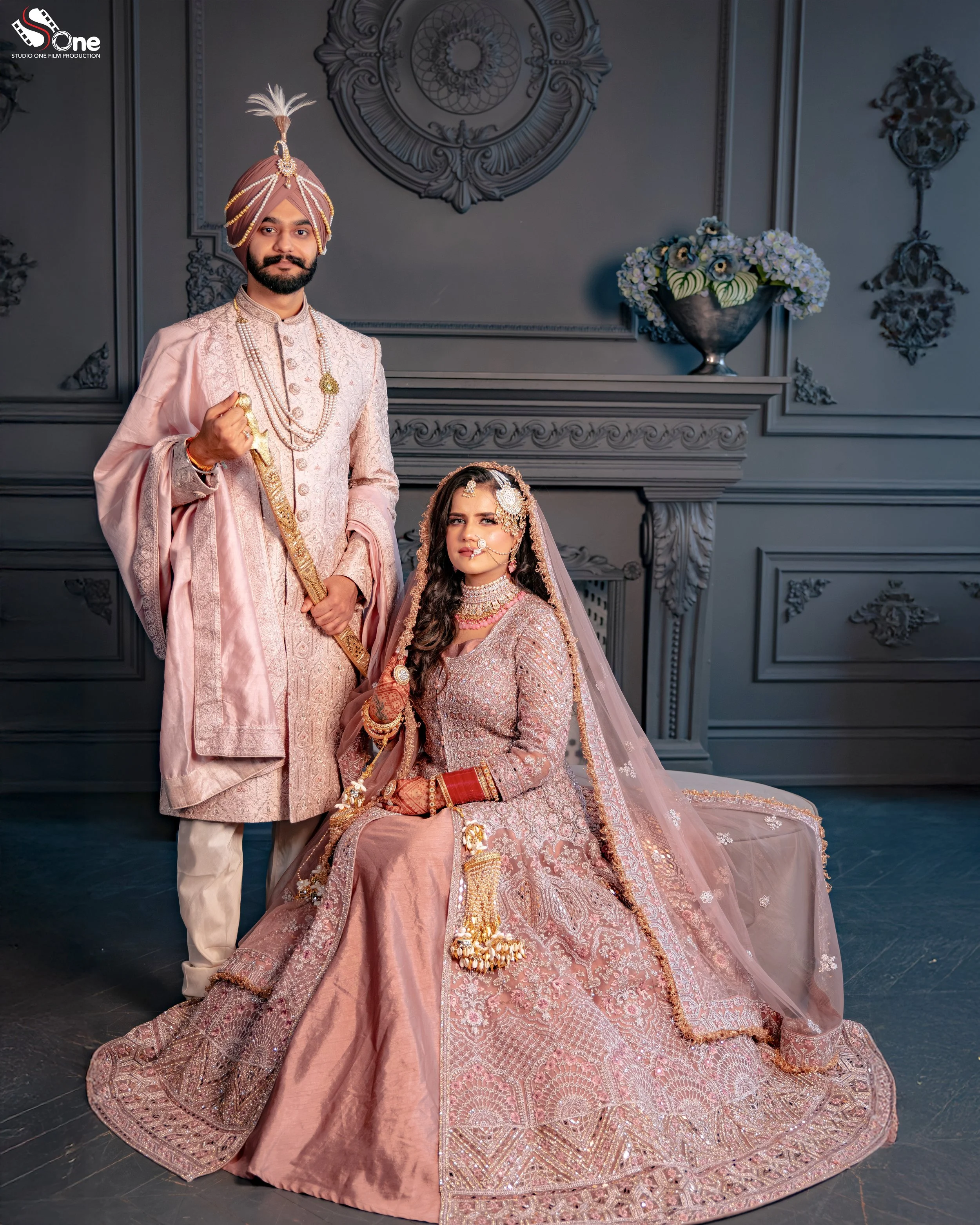 A couple dressed in traditional Indian wedding attire, the man in a pink sherwani and turban, the woman in an ornate pink bridal lehenga with jewelry, sitting and standing in a decorated indoor setting.