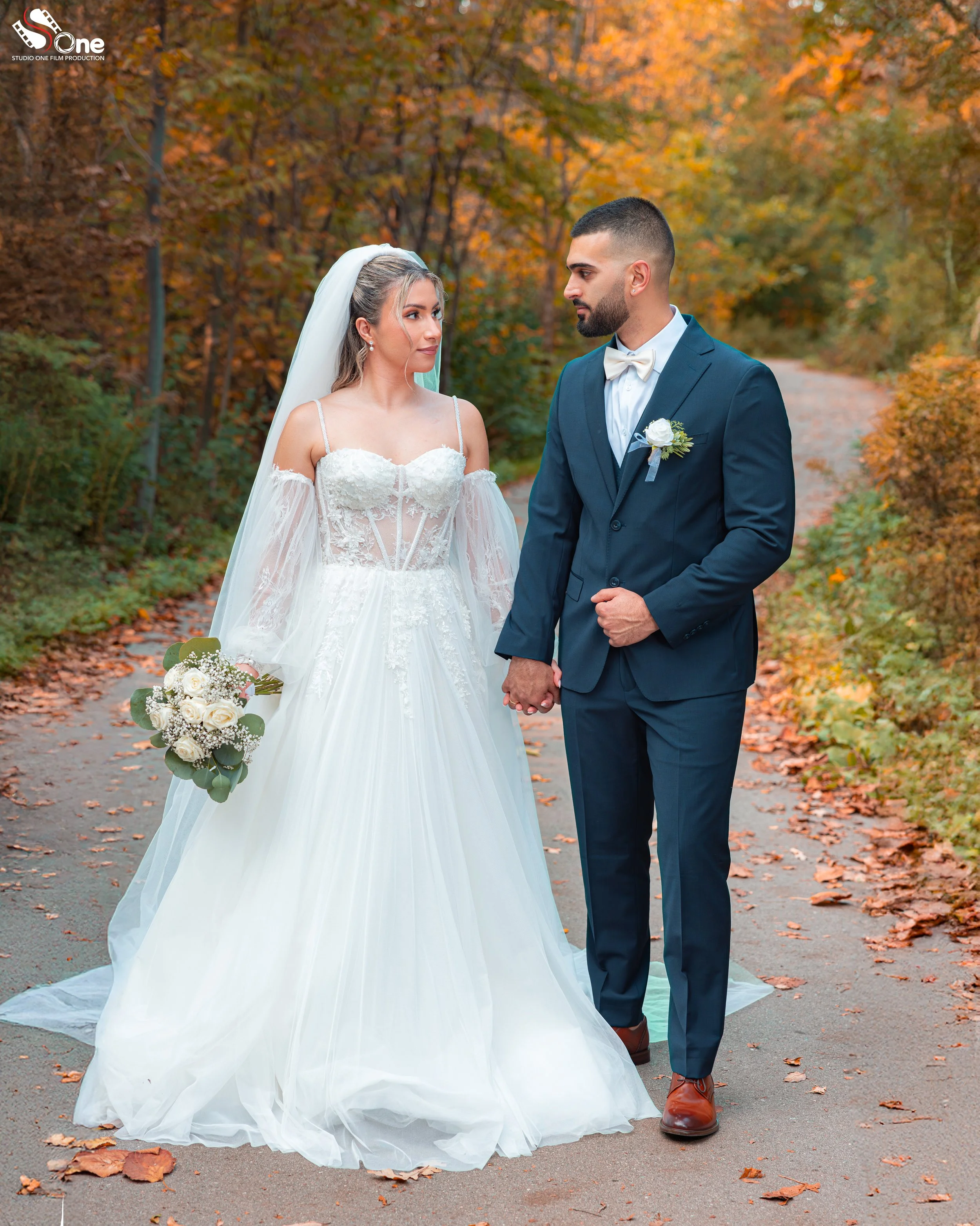 A bride in a white wedding gown holding a bouquet and a groom in a navy suit with a white bow tie standing on a leaf-covered path in an autumnal forest, holding hands and gazing at each other.