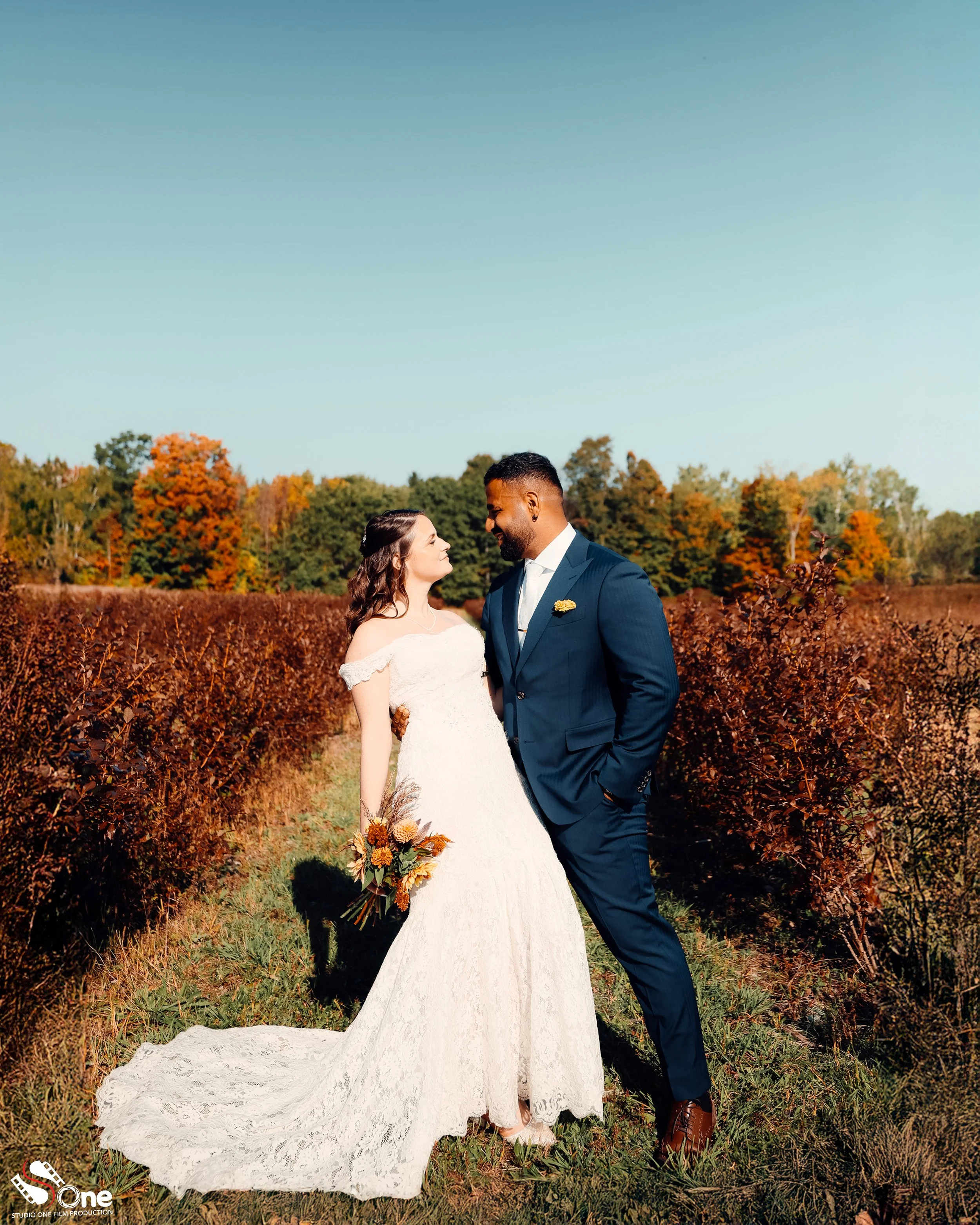 A bride in a white lace wedding gown holding a bouquet stands next to a groom in a blue suit in an autumnal field, smiling at each other with colorful trees in the background.