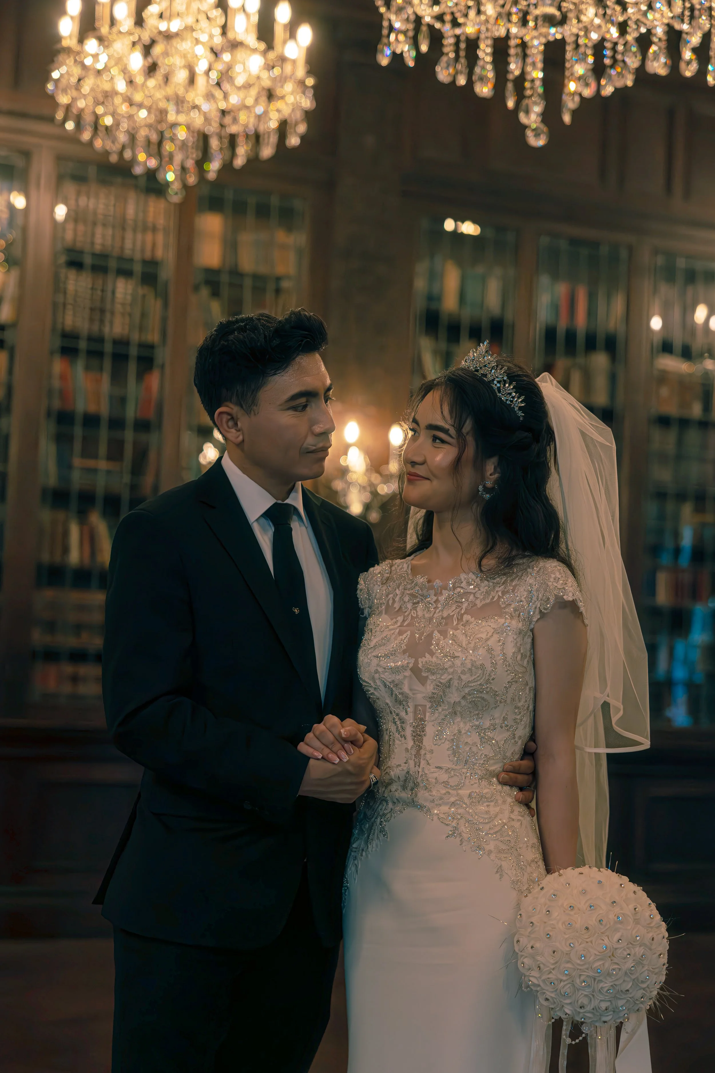 A bride and groom on their wedding day standing close in a warmly lit room with chandeliers and bookshelves in the background. The bride wears a white lace wedding dress and tiara, holding a bouquet of white roses. The groom wears a black suit and tie, holding her hand gently.