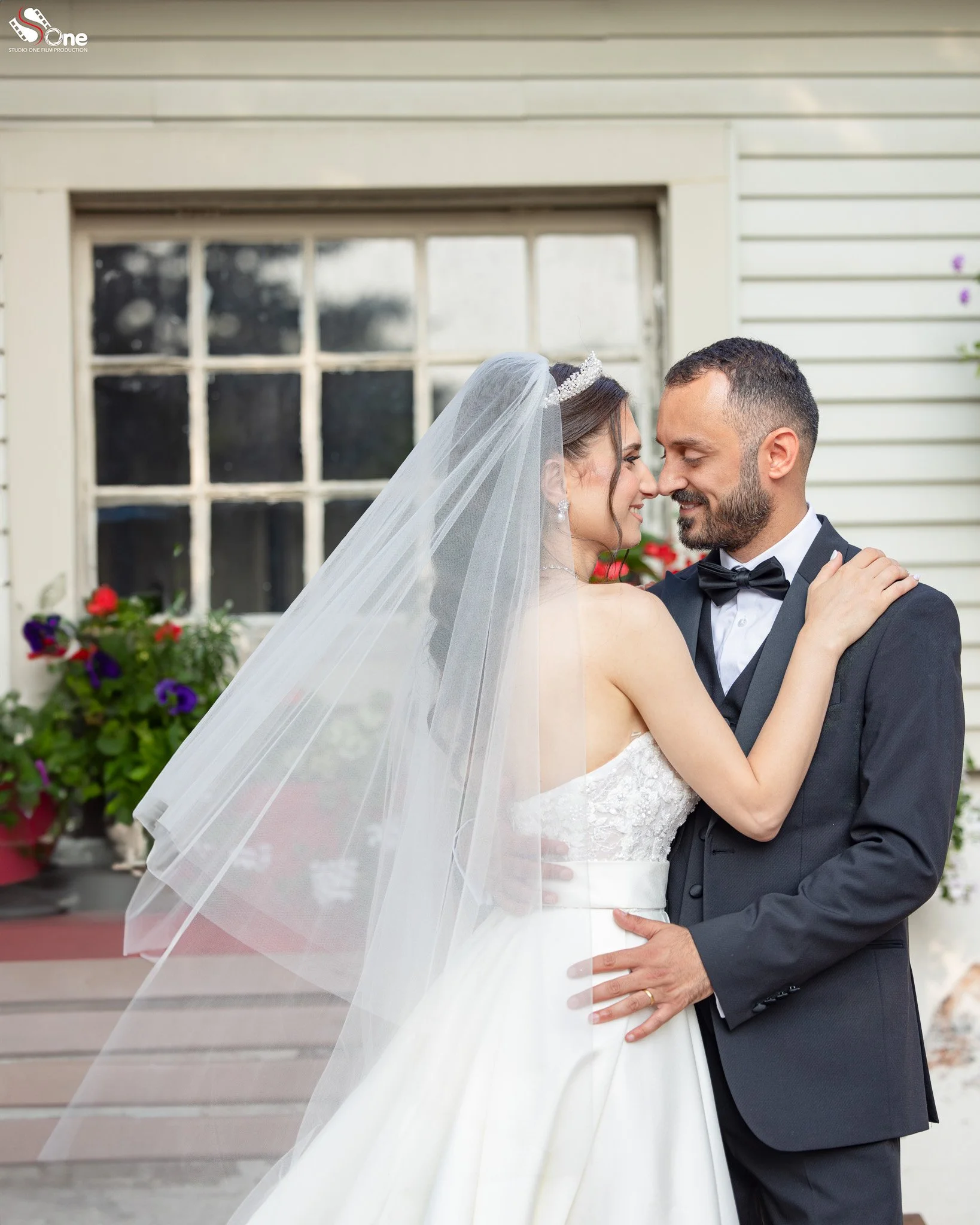 A bride and groom embrace outside a house, with the bride wearing a veil and wedding dress, and the groom in a black tuxedo, both smiling with their noses touching.