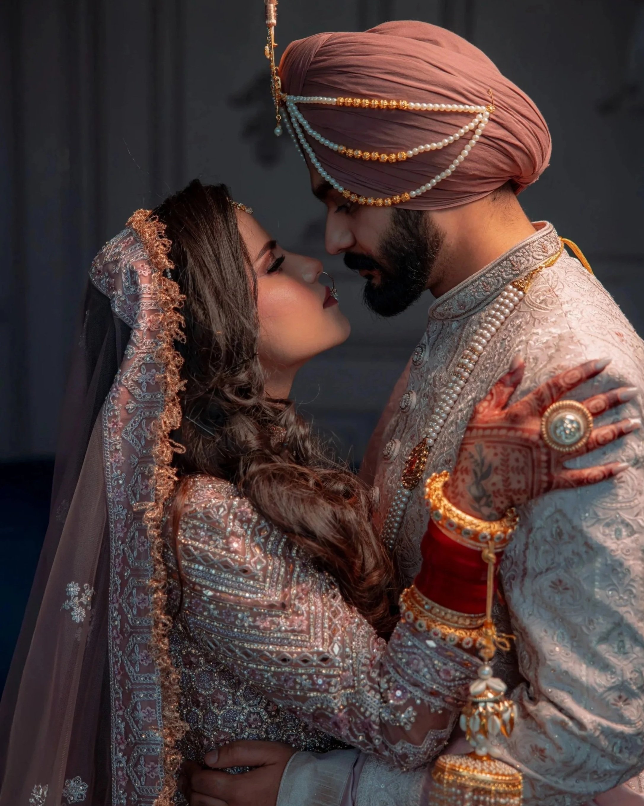 A bride and groom in traditional Indian wedding attire embrace closely, looking into each other's eyes indoors with soft lighting.