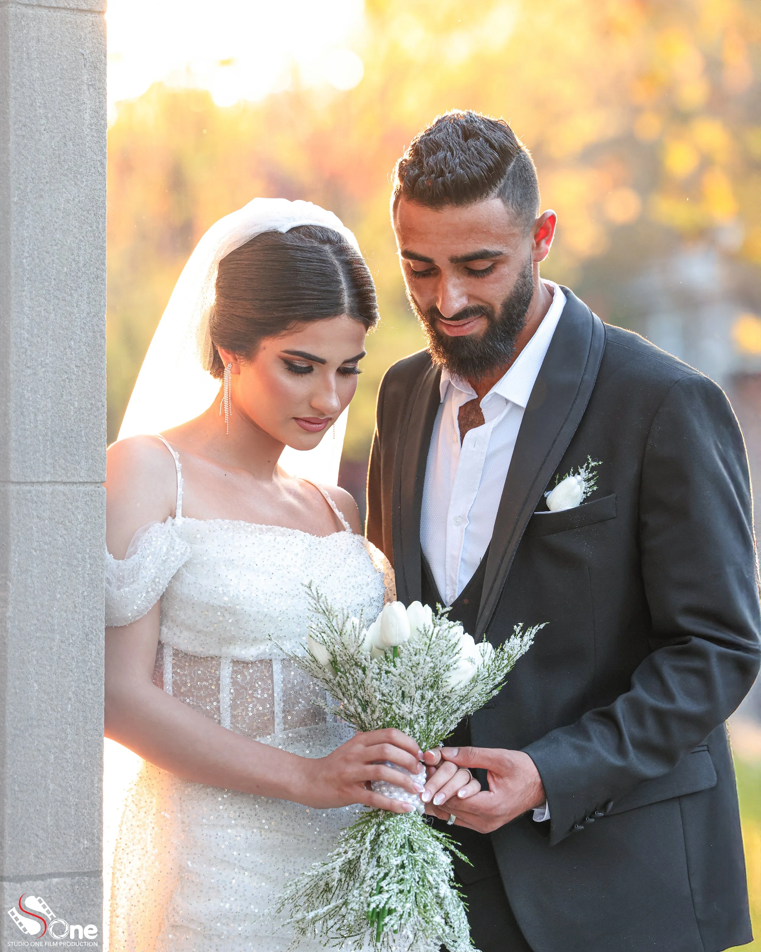 A bride and groom are standing outdoors during golden hour, with the bride holding a bouquet of white flowers and the groom gently holding her hand, both looking down.