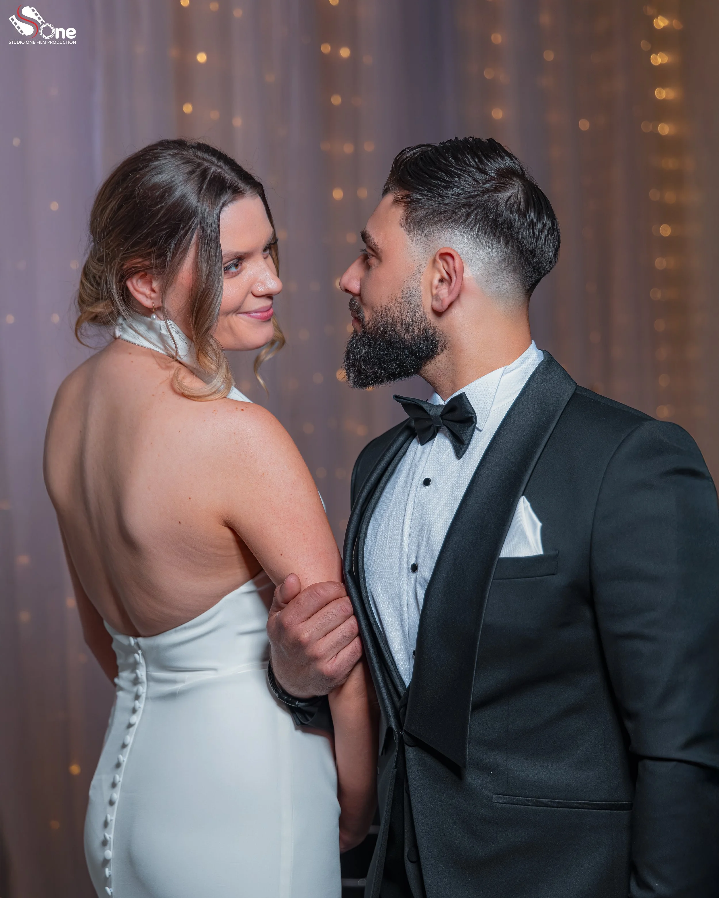 A couple in formal attire facing each other, the woman in a white dress with button details and the man in a black tuxedo with a bow tie, holding hands, and looking into each other's eyes at a wedding or formal event.