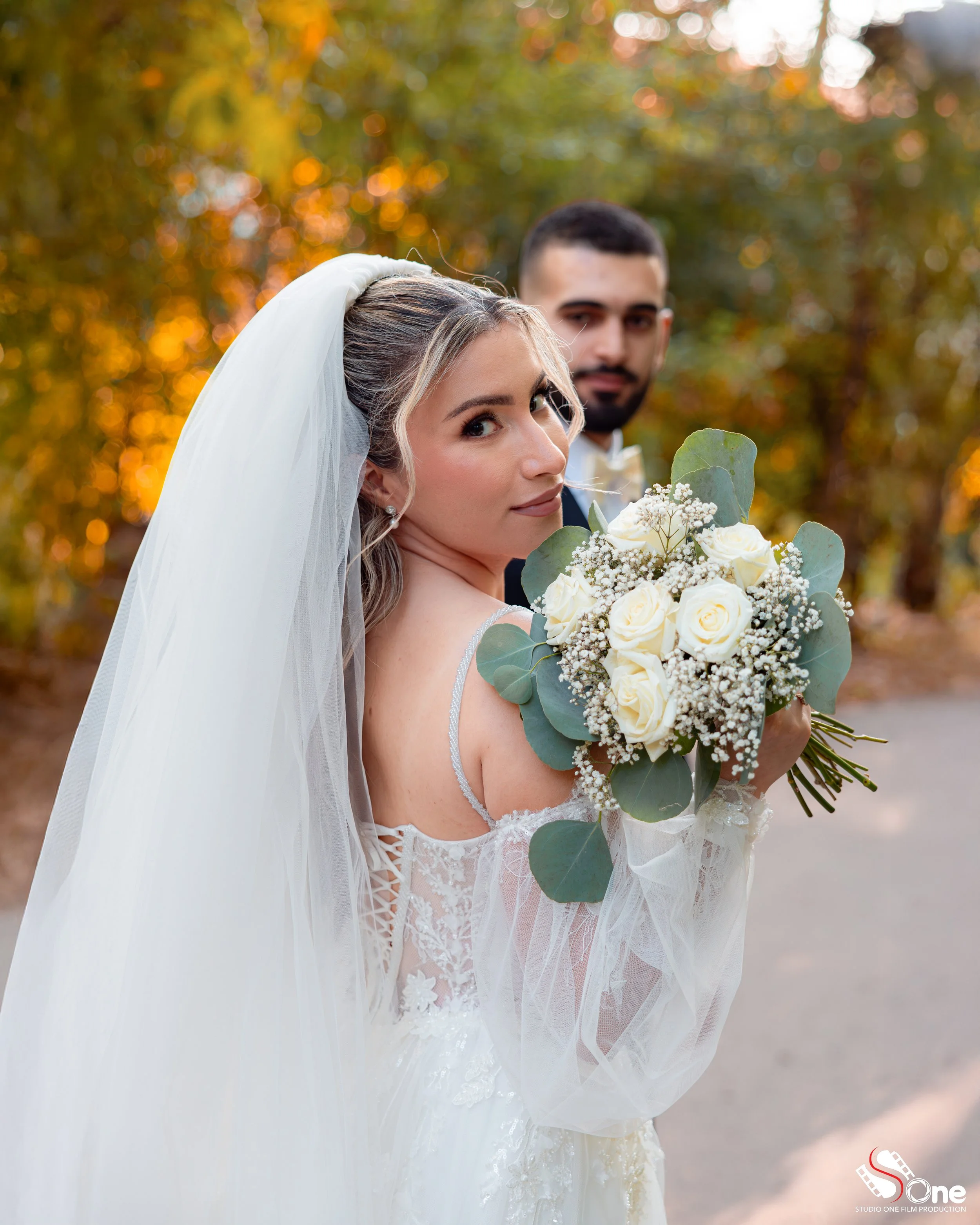 A bride in a white wedding dress with a veil holding a bouquet of white roses and greenery, standing outdoors with an autumnal background. A groom in formal attire stands behind her.
