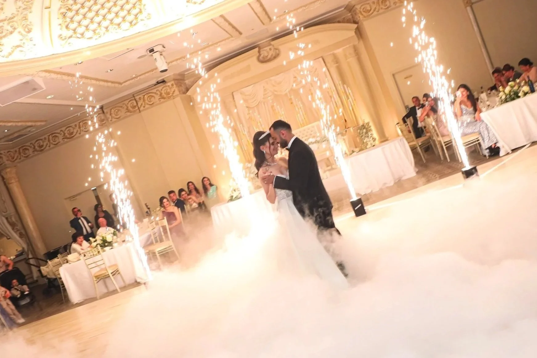 A bride and groom dancing in a decorated wedding hall surrounded by sparklers and fog, with guests seated at tables watching.