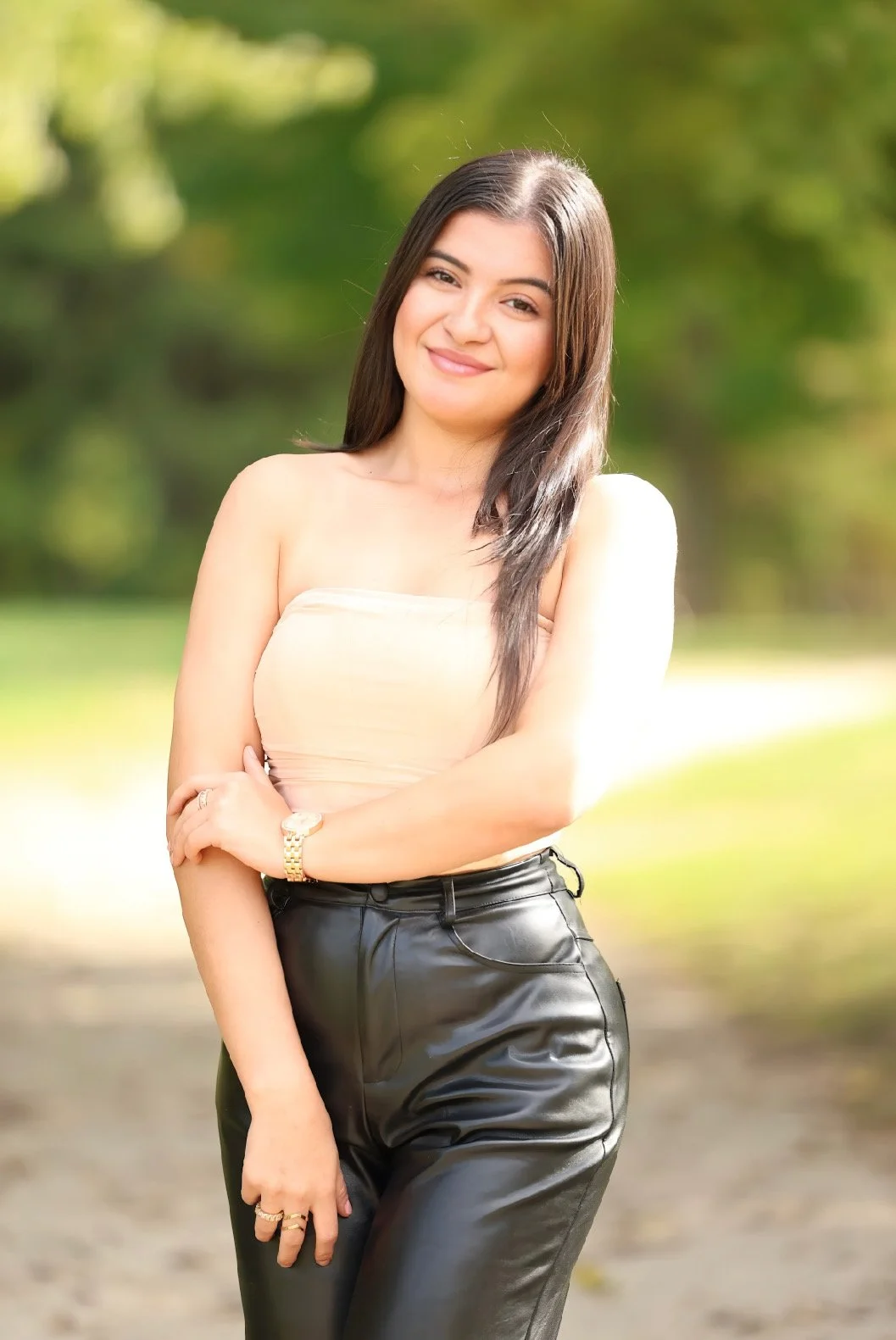 A young woman with long dark hair smiling outdoors in a park, wearing a strapless cream top and black leather pants, standing on a pathway with green trees in the background.