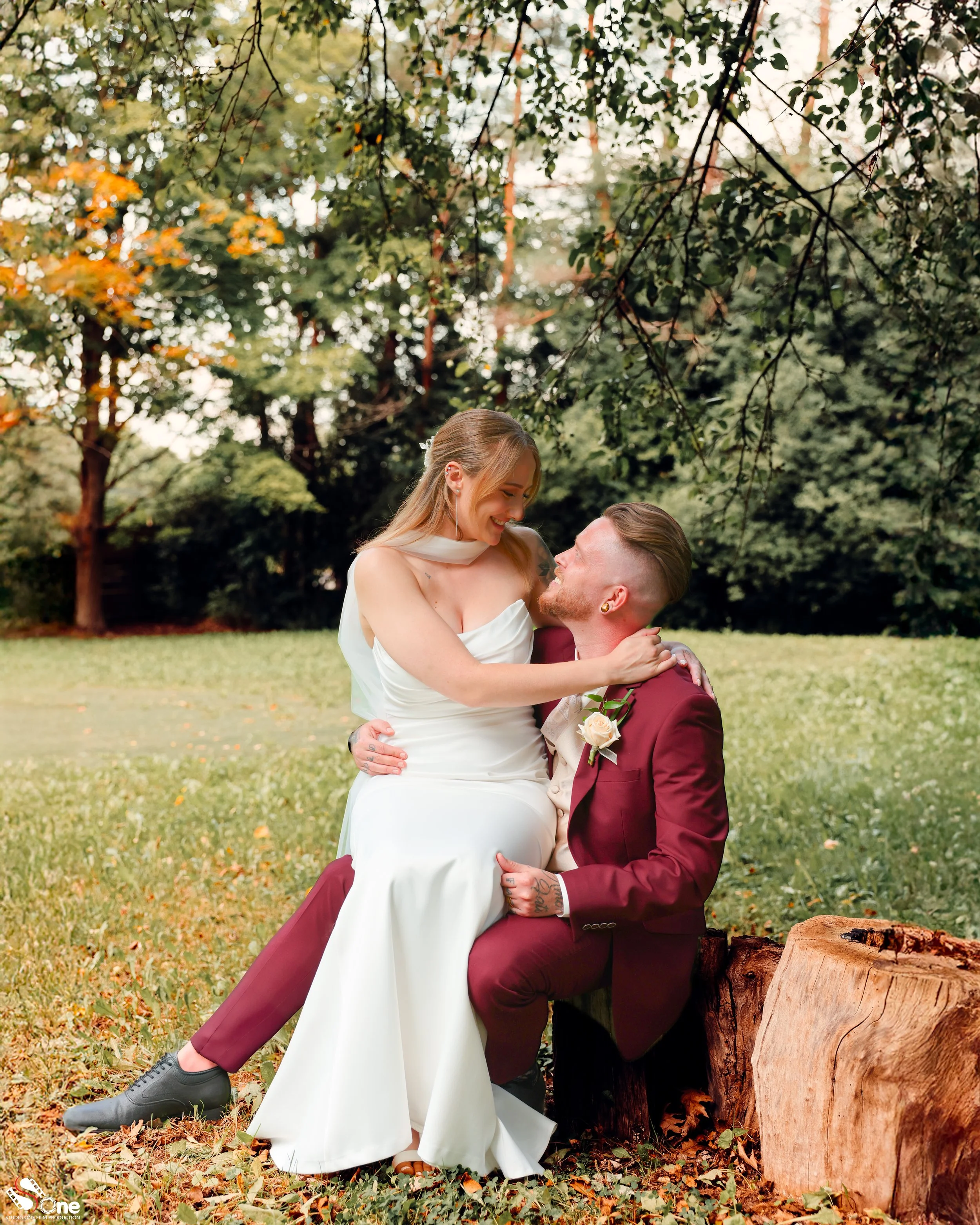 A bride and groom sitting on a fallen log in a park, the bride wearing a white wedding dress and the groom in a burgundy suit, sharing a joyful moment surrounded by green trees.