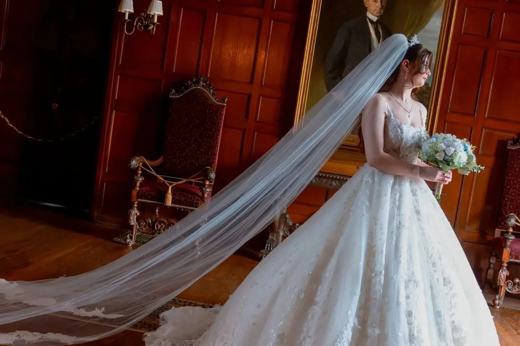 A bride in a wedding gown holding a bouquet of white and blue flowers, standing in a room with wood-paneled walls, a large portrait painting, and red velvet chairs.