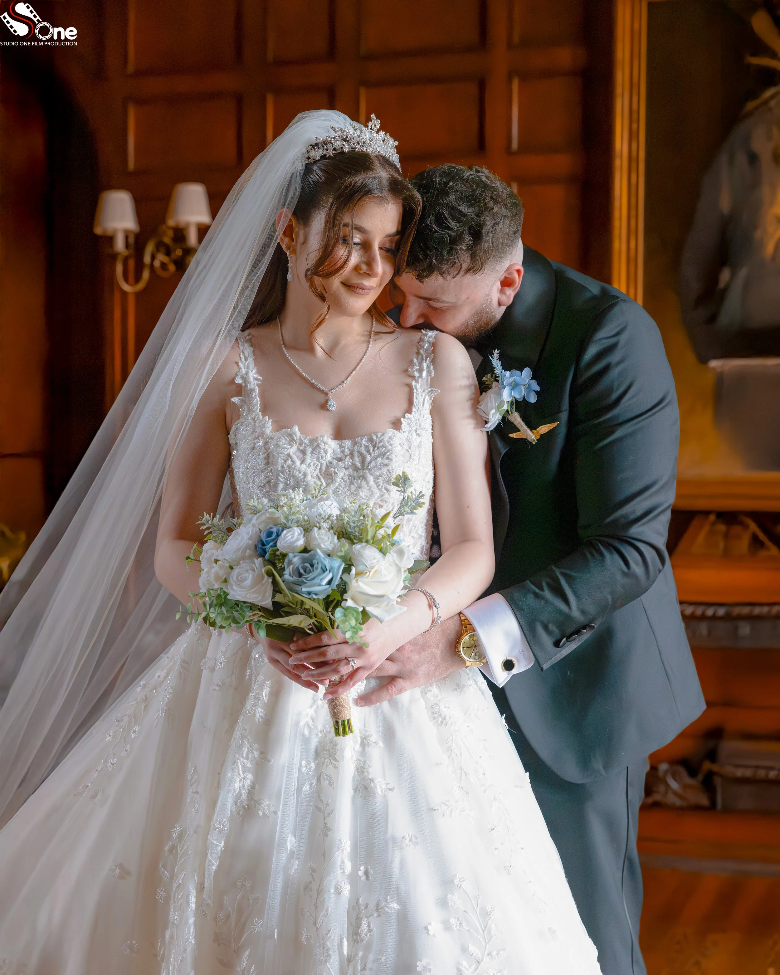 A bride and groom sharing a tender moment indoors. The bride is holding a bouquet of white and blue flowers, wearing a white wedding gown with lace detailing, and a veil. The groom is wearing a black suit with a white shirt and boutonniere, leaning in close to the bride.