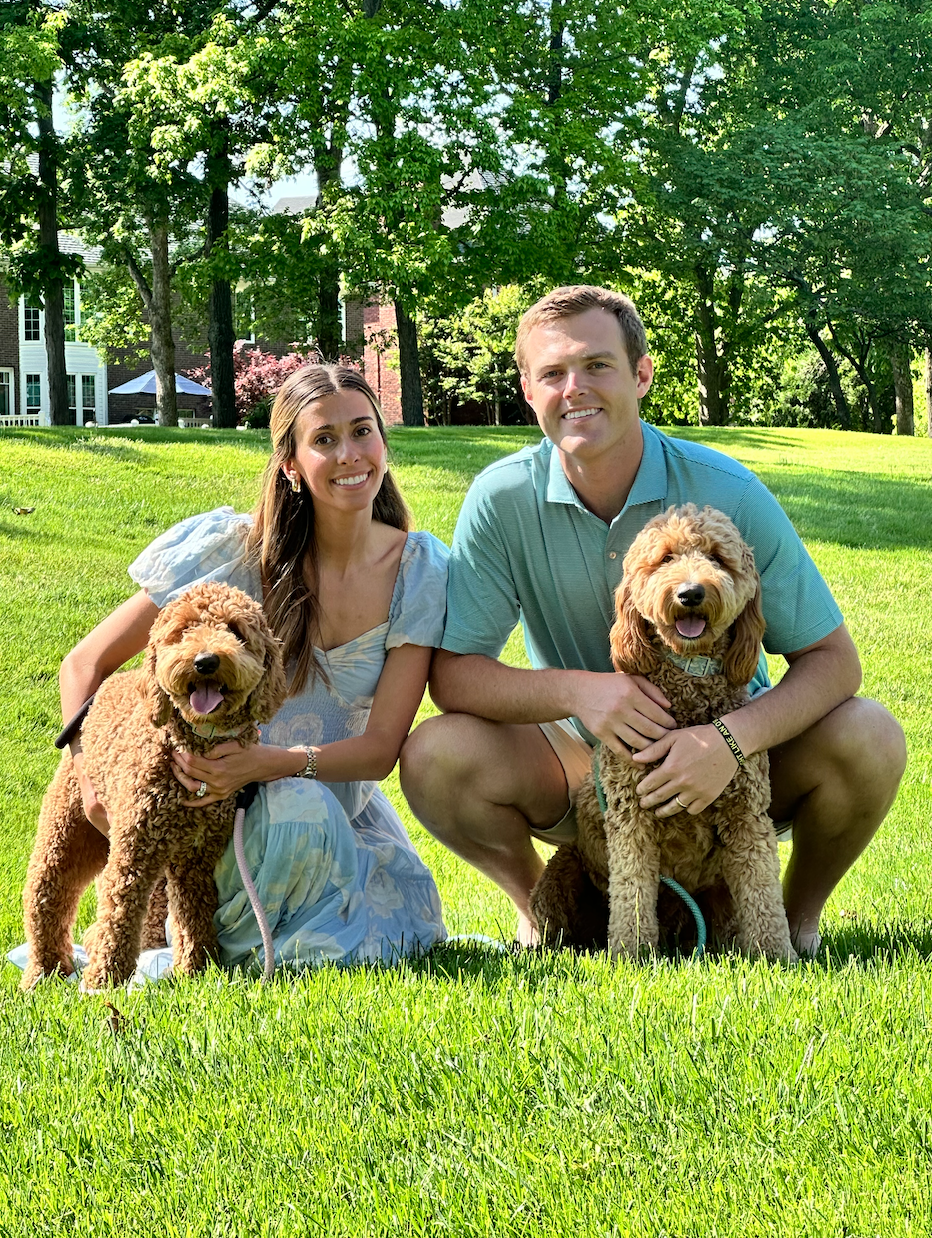 A smiling couple with two brown poodle dogs sitting on green grass in a park with trees and houses in the background.