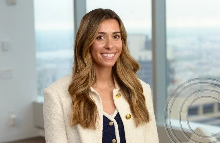A woman with long wavy light brown hair smiling, wearing a cream-colored blazer with gold buttons and a navy top, standing in an office with large windows showing a cityscape in the background.