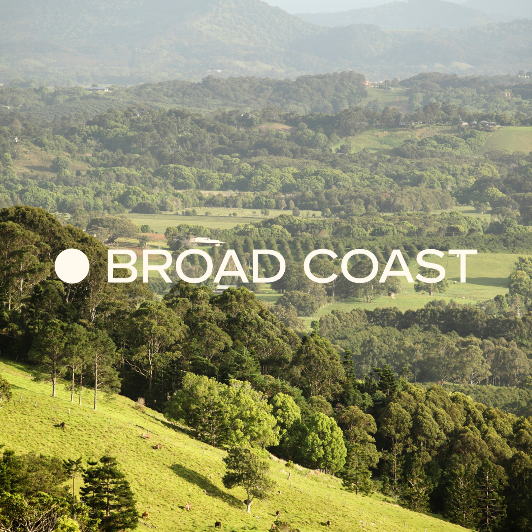 Lush green rolling hills with trees, grazing cows, and distant mountain ranges in the background, overlaid with 'BROAD COAST' text.