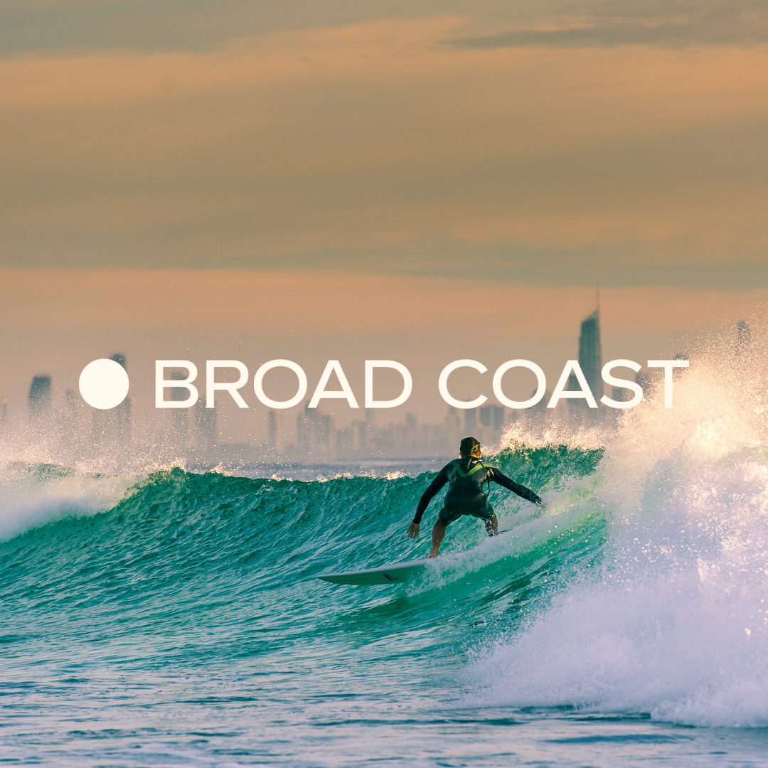 Surfer riding a wave in the ocean with a city skyline in the background during sunset or sunrise.