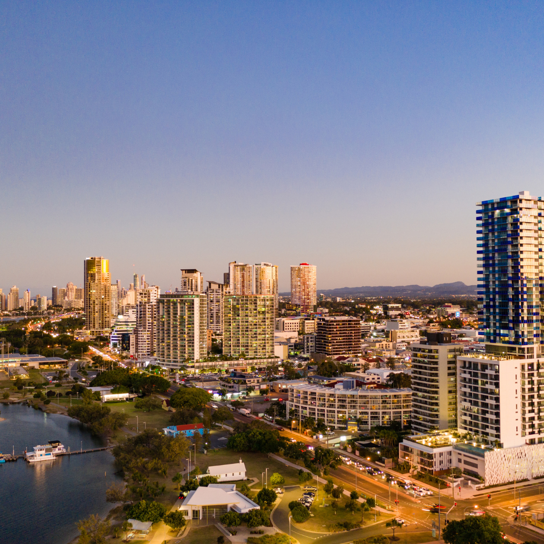 City skyline at sunset with modern high-rise buildings, a water body, and a road with moving cars in the foreground.