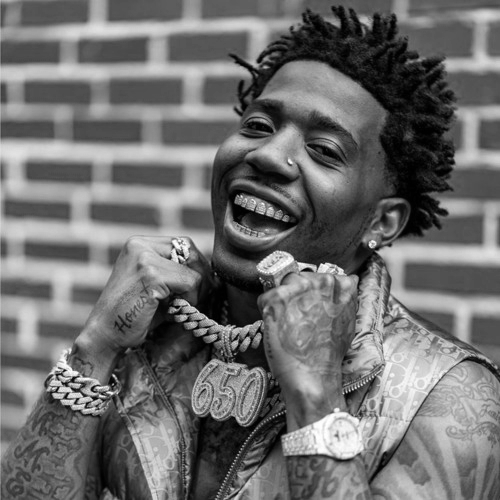 Young man with dreadlocks posing against a brick wall, smiling and showing gold teeth, wearing jewelry including rings, bracelets, a chain with a large pendant, and a watch.