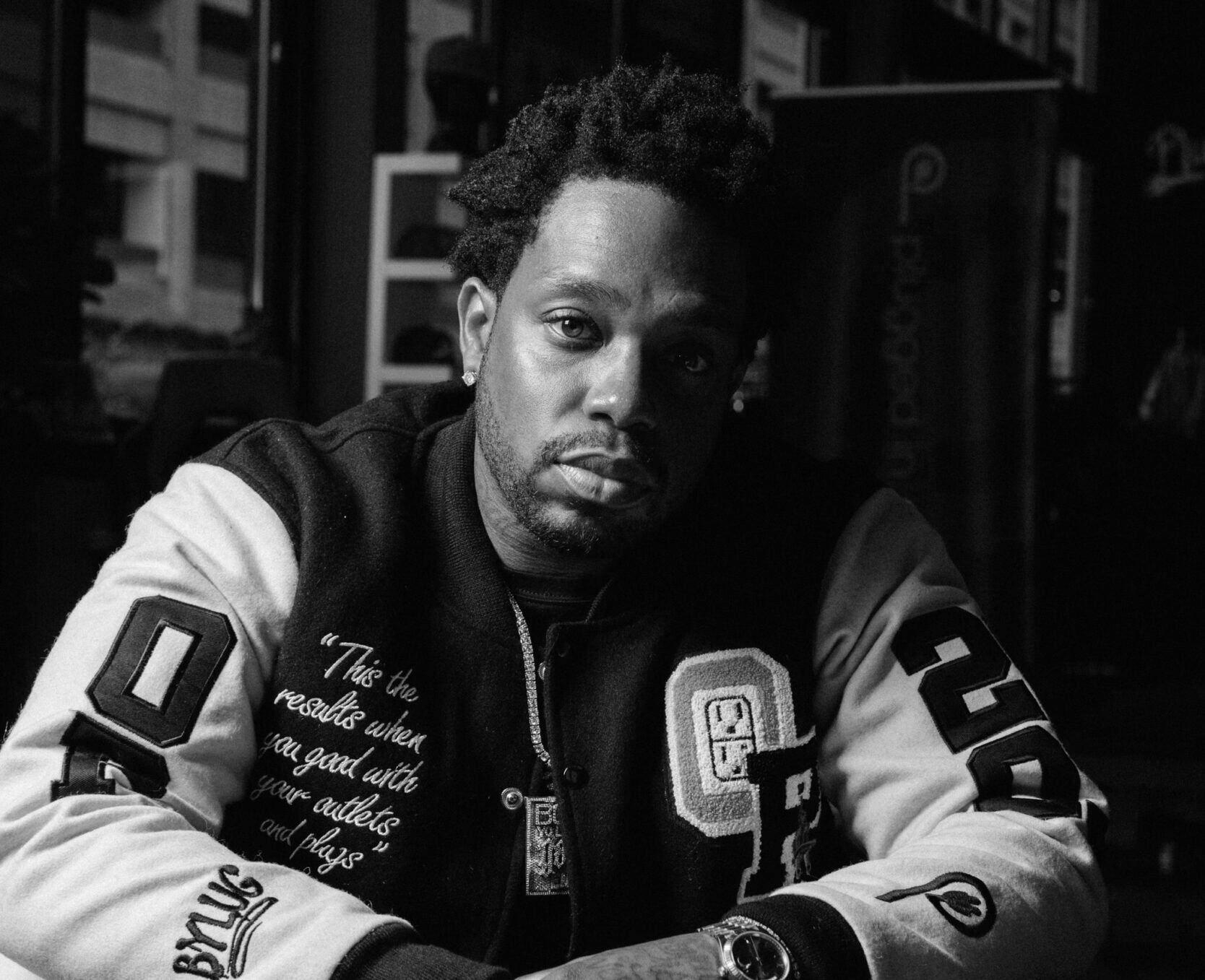 A black and white photo of a man with short, curly hair, wearing a varsity jacket with patches and embroidery, sitting indoors with a serious expression.
