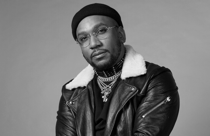 Black and white portrait of a man wearing glasses, a black beanie, a leather jacket with a shearling collar, and multiple layered necklaces.