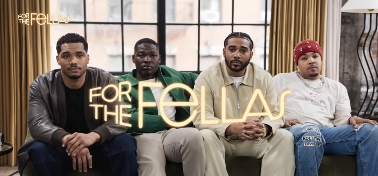 Four men sitting on a couch in front of a large window with golden curtains, with a neon sign reading 'For the Fellas' in front of them.
