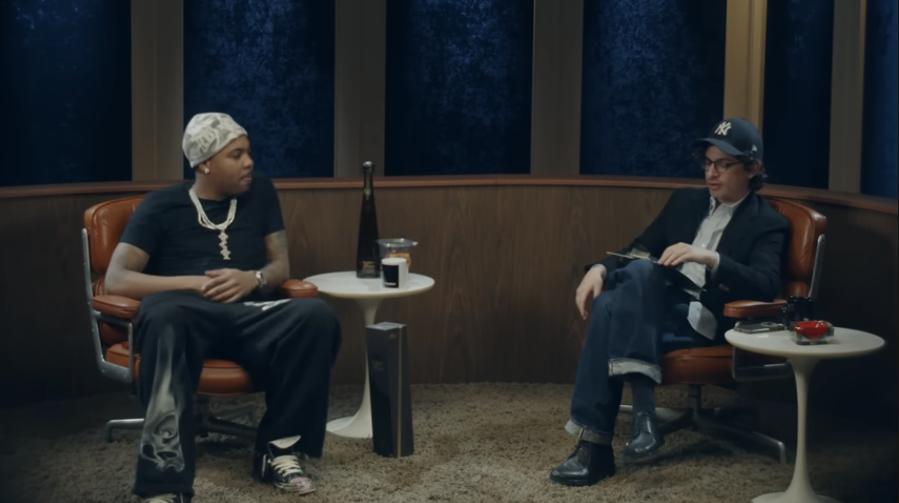 Two men sit in a dimly lit room, having a conversation. One man wears a black shirt, baggy pants, a white bandana, and jewelry. The other man wears a suit, glasses, a New York Yankees cap, and jeans. There are small tables with drinks and accessories between and beside them.