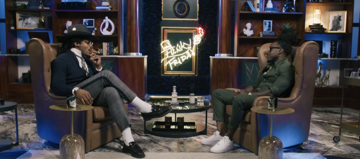 Two men sitting in armchairs facing each other during an interview or conversation in a studio decorated with bookshelves, artwork, and a neon sign that reads 'Funky Friday'.