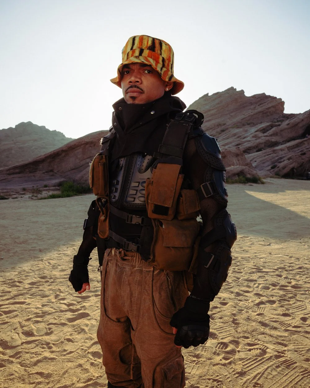 A man standing outdoors in a desert landscape with rock formations in the background. He is wearing a patterned bucket hat, black tactical gear, and brown cargo pants.
