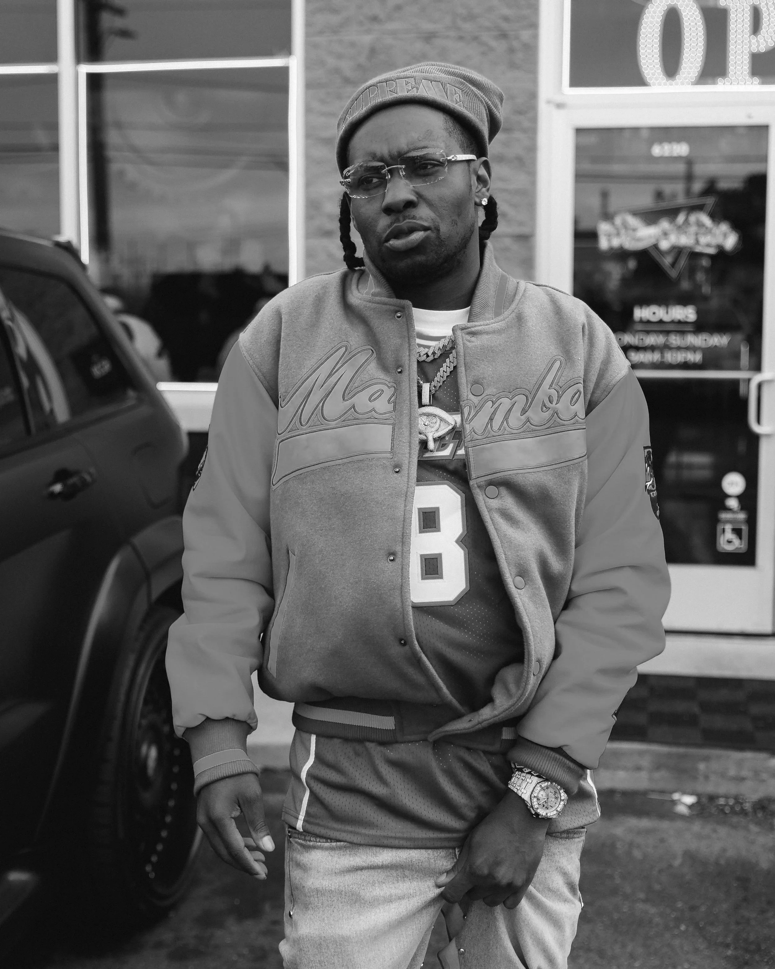 A man standing outside a store, wearing glasses, a baseball cap, a varsity jacket, a basketball jersey, and jewelry, with a car parked nearby.