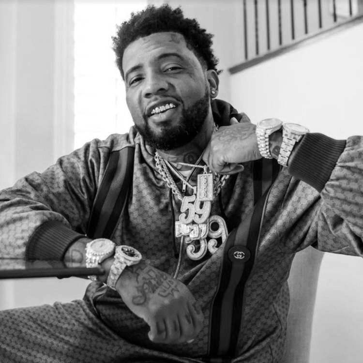 Black and white photo of a man with a beard and curly hair, smiling and resting his chin on his hand. He is wearing multiple jewelry pieces, including a large chain necklace, watches, and rings, and is dressed in a patterned jacket. He appears to be in a well-lit indoor setting.