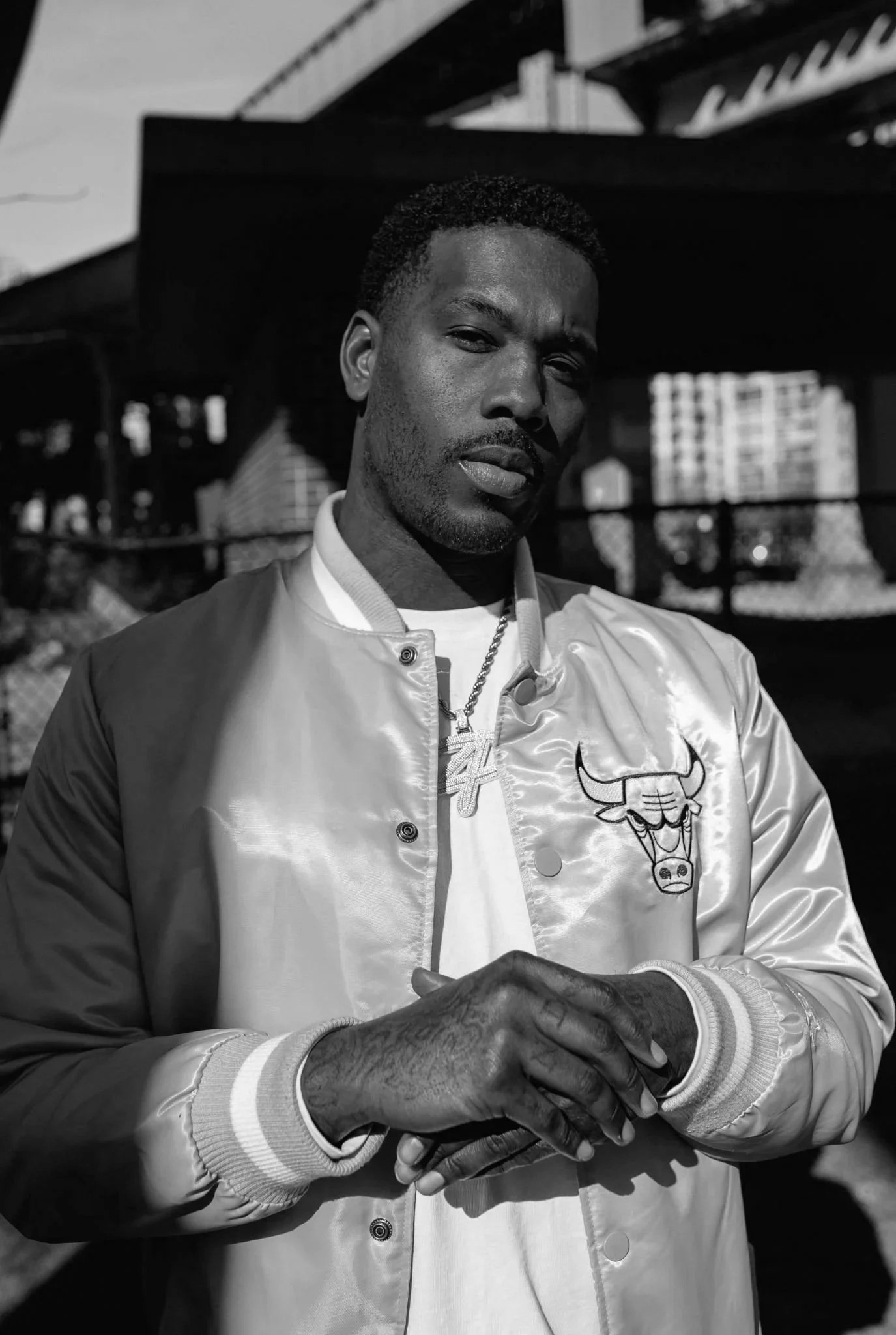 Black and white photo of a man with short curly hair and a beard, wearing a shiny bomber jacket with a Chicago Bulls logo, a white shirt, a chain necklace, and clasped hands in front of him, standing outdoors in an urban setting with buildings and a bridge in the background.