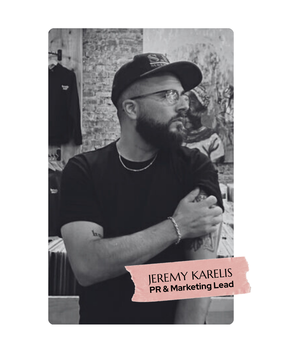 Black and white photo of Jeremy Karelis, a PR and Marketing Lead, with a beard, glasses, wearing a hat, a black t-shirt, and a chain necklace, standing in a store with exposed brick wall and clothing in the background, with a pink overlay showing his name and job title.