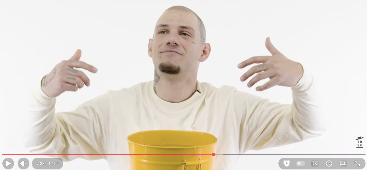 A man with short hair, a goatee, and tattoos on his neck and hands, wearing a light-colored long-sleeve shirt, poses in front of a white background with a yellow bucket in front of him. He is touching his head and shoulders with a confident expression.