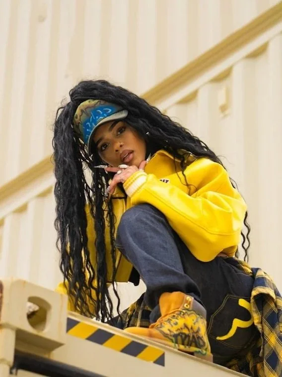 Black women crouched, posing and looking at the camera. Dressed in a white windbreaker, gray sweatpants, beige work boots and 
