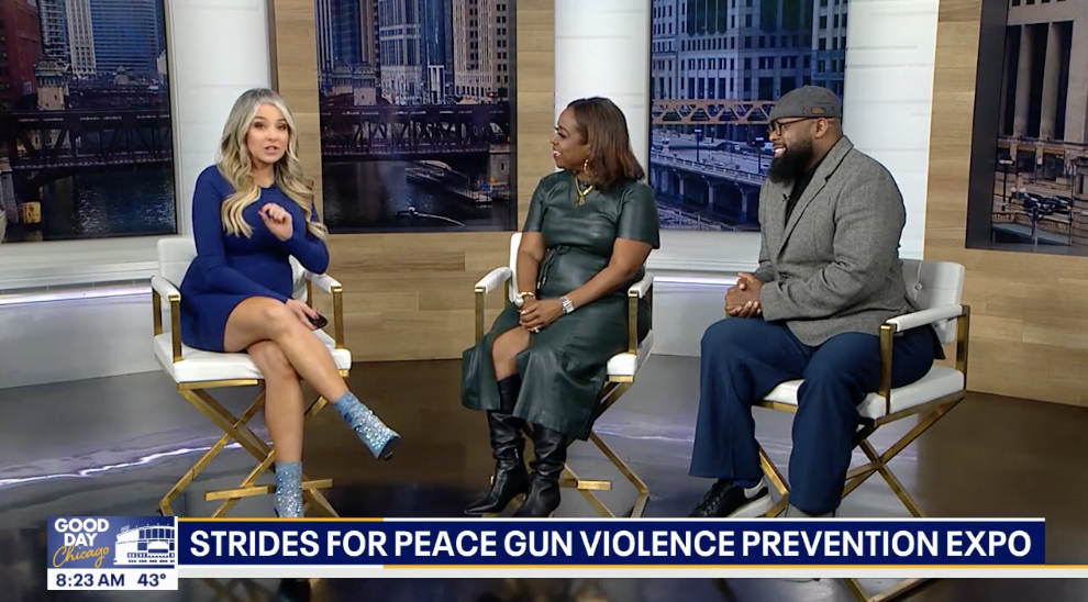 Three people sit on a TV set during an interview. The woman on the left is talking, wearing a blue dress and sparkly blue boots. The woman in the middle wears a dark green dress, and the man on the right is dressed in a gray coat, blue pants, and a gray cap. Behind them are large cityscape screens. A news caption at the bottom reads 'Strides for Peace Gun Violence Prevention Expo,' with the time 8:23 AM and temperature 43°F displayed.