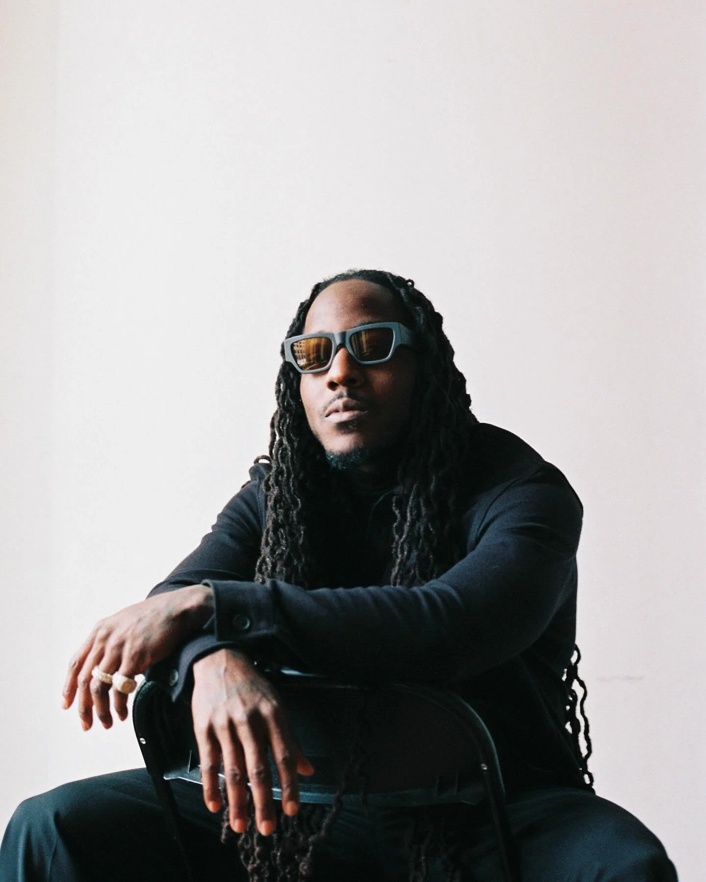 A man with long dreadlocks wearing black clothing and sunglasses, sitting on a chair against a plain white wall.