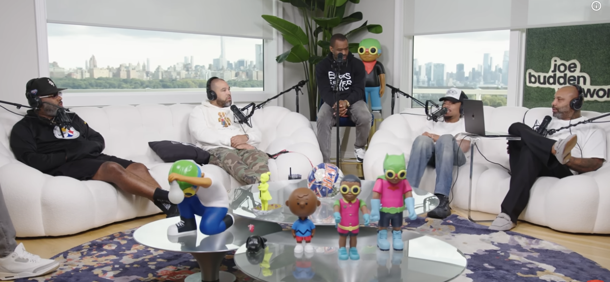 A podcast recording session with five individuals seated on white couches, each wearing headphones and speaking into microphones, in a room with large windows and a city skyline view. The table in front displays colorful toys, including characters from the animated series 'Rugrats' and other figurines.