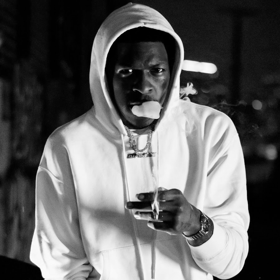 A young man with dark skin wearing a white hoodie, holding a lit cigarette or similar object in one hand, with smoke coming from his mouth and nose, in a dimly lit environment.